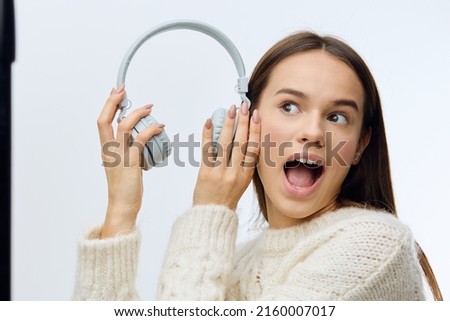a funny, emotional woman stands putting headphones to her ear in a knitted white sweater and opens her mouth wide with delight. Horizontal Studio Photography