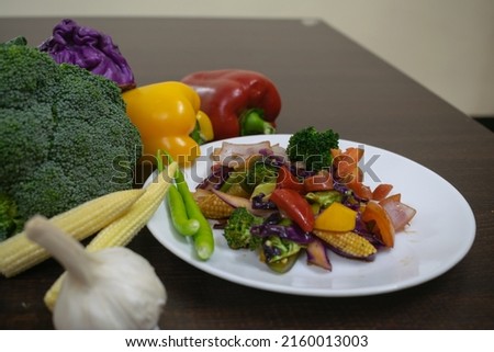 Picture of sautéed salad on a white plate along with healthy raw boiled exotic vegetables. baby corn, capsicum, bell pepper, broccoli, garlic, chilly, cabbage, tomato, onion, carrot on wooden table.