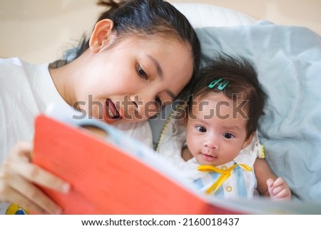 Smiling young Asian mother sit on the bed with little infant toddler child, happy biracial mom relax have fun read tale book with small baby girl at home, motherhood, childcare concept Royalty-Free Stock Photo #2160018437