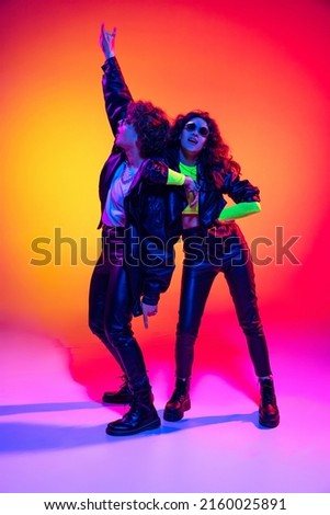 Excited young man and girl in black leather outfits having fun, gesturing, shouting and jumping on yellow-red background in neon light. Concept of style, music, fashion and youth