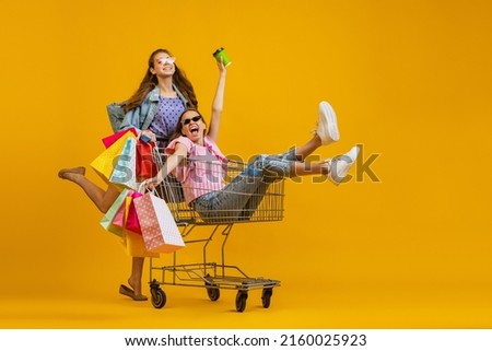 Ready for big sales. Beautiful young astonished girls go shopping with shopping cart isolated on bright yellow background. Concept of sales, black friday, discount, emotions. Copy space for ad, text Royalty-Free Stock Photo #2160025923