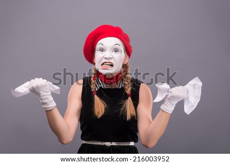 Waist-up Portrait of female mime with red hat and white face angry crumpling a paper isolated on grey background with copy place