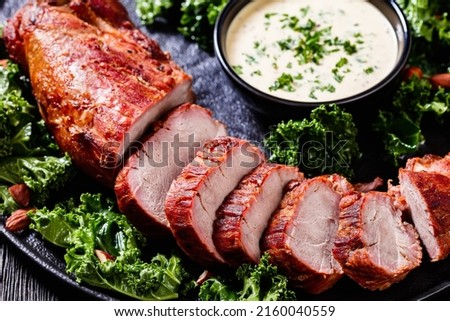 close-up of Roast Pork Tenderloin with Creamy Mustard Tarragon Sauce on black plate with fresh kale and roast almond salad on dark wooden table, horizontal view from above