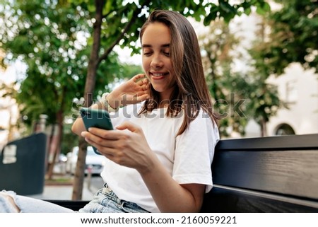 Alluring woman with loose hair in white short is touching her hair and looking at smartphone while sitting on city bench and waiting for friends . High quality photo