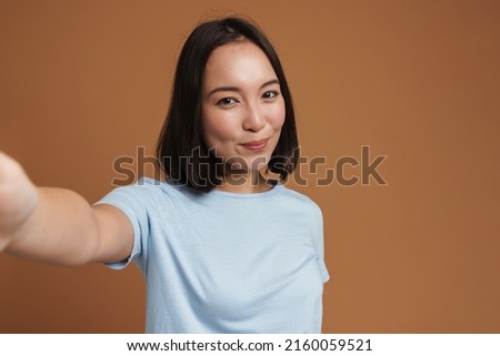 Young asian woman laughing while taking selfie photo isolated over beige background