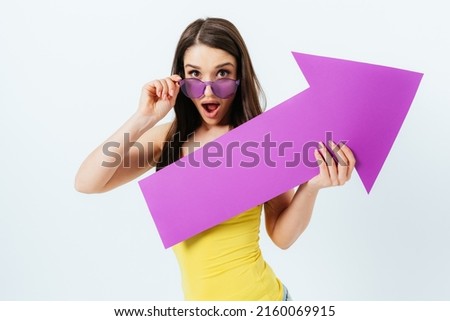 Portrait shocked brunette girl wearing summer outfit and sunglasses pointing away with big arrow while standing over white background