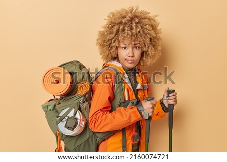 Sideways shot of serious curly haired woman trekking in mountains poses with walking poles carries rucksack dressed in orange jacket isolated over beige background. People and hiking concept