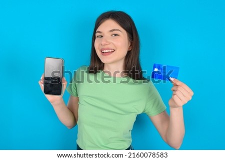 Young Caucasian woman wearing green T-shirt over blue background   opened bank account, holding smartphone and credit card, smiling, recommend use online shopping application