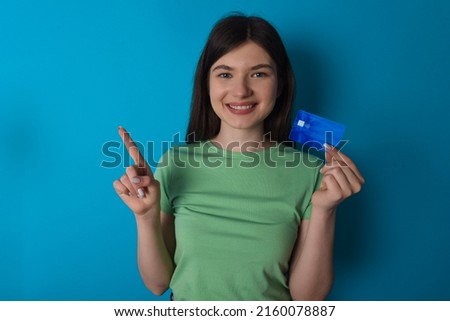 Young Caucasian woman wearing green T-shirt over blue background showing debit card pointing finger empty space 