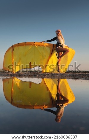 Attractive stylish young caucasian woman in cap sunglasses and kitesurfer outfit standing next to sandy shore next to her kite at sunset reflecting in the water