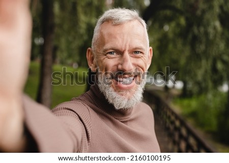 Smiling caucasian mature man looking at the camera, taking selfie on smart phone, having video call, vlogging blogging outdoors in park.