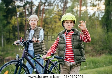 Happy grandmother with her grandson on their bikes walking on wooden forest. School age boy shows sign ok.