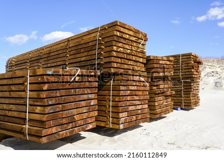 Stacked impregnated brown pine wood boards by the sea on a sandy beach, blue sky background Royalty-Free Stock Photo #2160112849