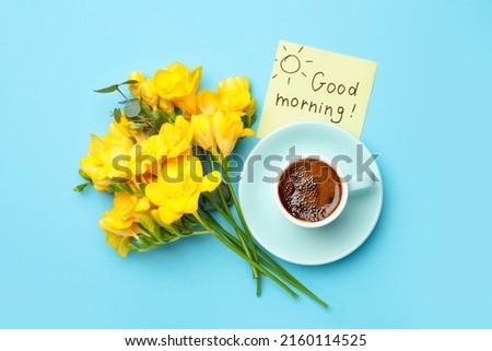 Cup of aromatic coffee, beautiful yellow freesias and Good Morning note on light blue background, flat lay