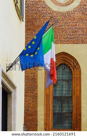 Flags of the European Union and Italy after the rain over an open doorway against the background of a red brick wall and a window of a gothic building, side view vertical orientation
