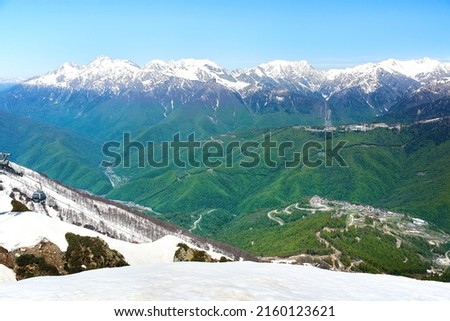View from Rosa Peak to the snow-capped mountain peaks and the lower station of the Rosa Dolina ski lift in the greenery of trees. Krasnaya Polyana, Sochi, spring.