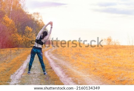 Young slim brunette in sportswear doing exercises and stretching on field against backdrop of dawn. Morning autumn workout in nature.Gymnastics and yoga is good way of exercise to stay fit and healthy