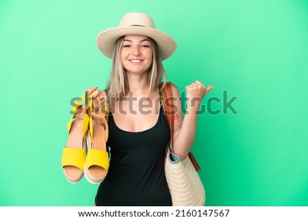 Young Romanian woman in swimsuit holding sandals for go to the beach isolated on green background pointing to the side to present a product