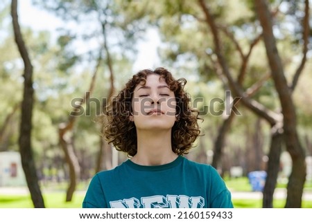 Young redhead woman wearing green tee standing on green city park, outdoors. Enjoying stress free mindful moment, doing yoga relaxation exercises, closed eyes, meditating near nature, taking breath.