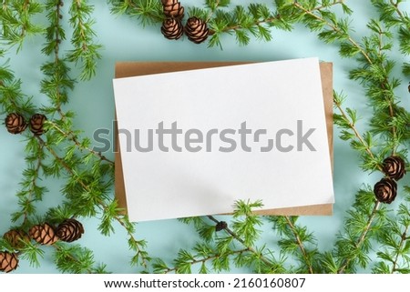 A mock-up of a kraft envelope with a blank sheet and a branch of green larch on a light blue background. Copy space. Flat lay, top view.