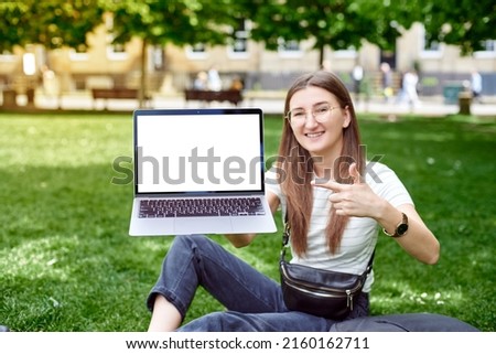 A young pretty woman holds a laptop in front of her and points her finger at it. Mockup of a laptop with a blank screen. A girl shows off a laptop on the street on a green background.
