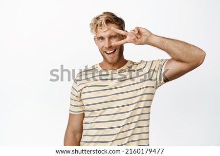 Portrait of blond stylish guy showing peace, v-sign gesture and smiling, winking at camera, standing over white background