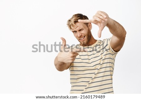 Portrait of creative blond guy picturing, taking good shot, searching for perfect angle with hand frame gesture, measuring something on eye, white background
