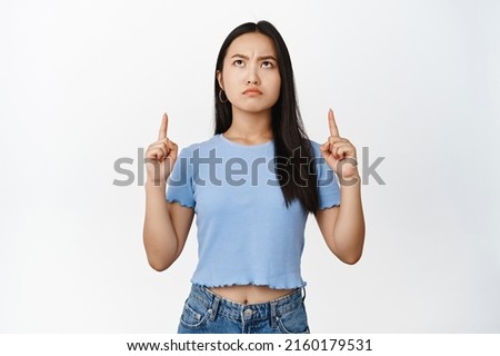 Moody asian girl pointing and looking up, frowning with angry face expression, standing in blue tshirt over white background