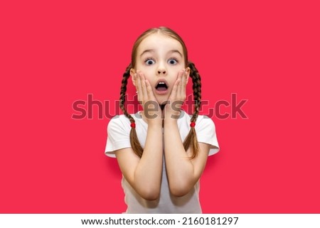 Girl child with pigtails on a red background with a surprised look holds on to her face. Shocked little girl with big eyes and open mouth. Child with a grimace of shock and surprise