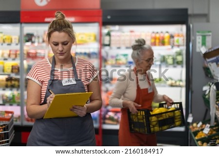 Young shop assistant in supermarket in vegetable shell, in bakcground is her colleague filling stock.