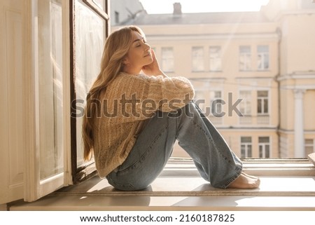 Cute caucasian adult lady with her eyes closed is basking in sun sitting on windowsill on warm day. Long-haired blonde wears casual clothes. Concept of enjoying moment Royalty-Free Stock Photo #2160187825