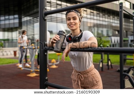 One caucasian woman taking a brake during outdoor training in the park outdoor gym resting on the bars with supplement shaker in hand drinking water or supplementation happy smile copy space Royalty-Free Stock Photo #2160190309