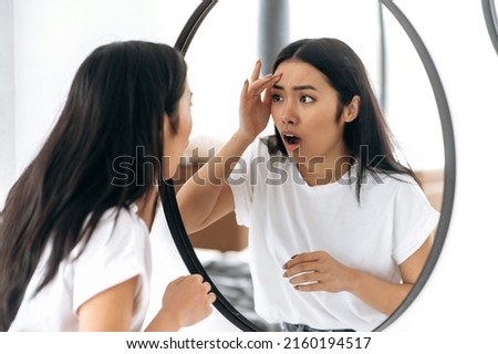 Amazed sad Chinese girl in a white t-shirt, standing in front of a mirror at home, looking at herself in the mirror in shock, saw a pimple or wrinkle, unpleasantly surprised, needs facial skin care Royalty-Free Stock Photo #2160194517
