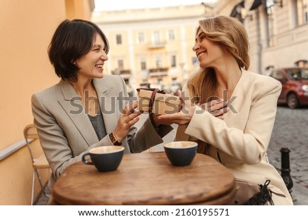 Happy caucasian adult lady on right receives holiday gift box from her friend on left. Women have nice time in street cafe with cup of coffee. People sincere emotions lifestyle concept.