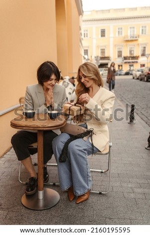 Full length image of two cute women sitting in street cafe with gift box in their hands. Blonde opens gift received from colleague. Holiday life concept