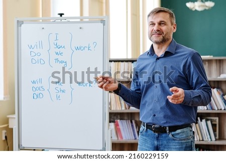 Portrait of mature Caucasian teacher wearing blue shirt and jeans standing next to whiteboard explaining English question grammar rules Royalty-Free Stock Photo #2160229159