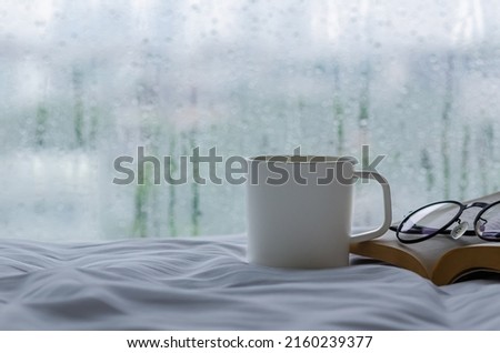 A cup of coffee with book and spectacles on bed in morning with rain drop on window. Stay home and relaxing concept. Royalty-Free Stock Photo #2160239377