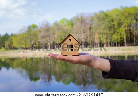 Miniature wooden house model in hand against a lake background in spring. Living in countryside.