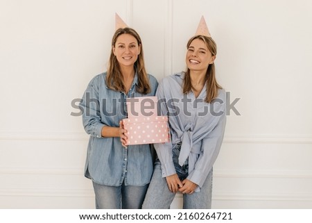 Image of two caucasian women smiling at camera holding gift boxes on white background. Blondes wear shirts, jeans and festive colacas. Holiday concept