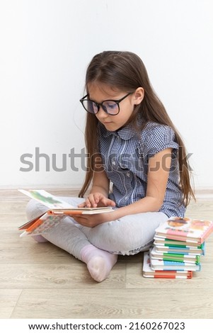A 7-year-old girl with glasses reads a book sitting on the floor. Children's education, learning concept.
