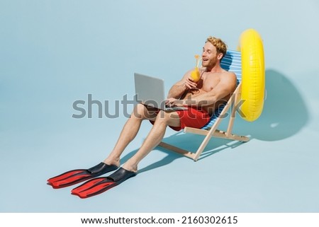 Full length young man 20s in red shorts swimsuit sit in deckchair inflatable ring hold cocktail use laprop pc computer work isolated on pastel blue background Summer vacation sea rest sun tan concept. Royalty-Free Stock Photo #2160302615