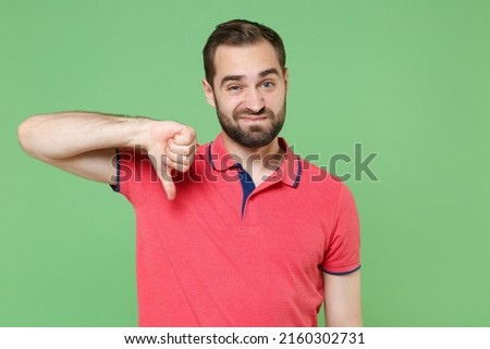 Displeased young bearded man 20s in casual red pink t-shirt posing isolated on green wall background studio portrait. People sincere emotions lifestyle concept. Mock up copy space. Showing thumb down