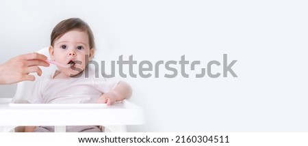 Banner copyspace baby girl eating blend mashed food sitting, on high chair, mother feeding child, hand with spoon for vegetable lunch, baby weaning, first solid food for young kid. Royalty-Free Stock Photo #2160304511