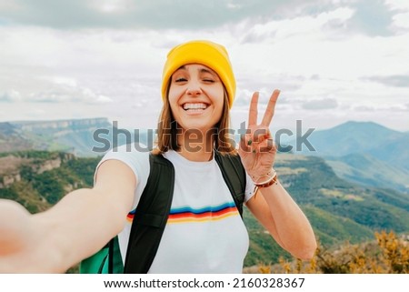 Young cheerful female backpacker with short brunette hair and yellow beanie, taking selfie with smartphone doing victory sign with fingers and winking an eye at camera on top rocky mountains.