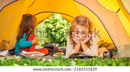 Small children, kids play,read book in orange tourist tent. Family trip, hike to nature. Backyard games, having fun. Outdoor recreation, activity. Self-assembly, setting up camping tent on lawn,grass. Royalty-Free Stock Photo #2160331839