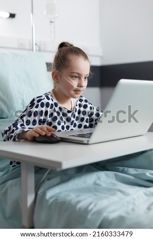 Smiling sick little girl playing games on computer laptop while in pediatric clinic recovery ward room. Happy ill little child enjoying video games on laptop while resting in hospital bed.