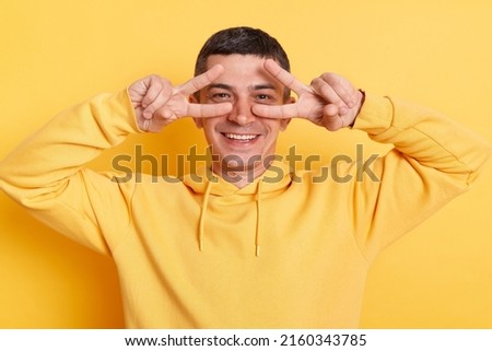 Smiling delighted man wearing casual style hoodie posing isolated over yellow background, standing with victory, peace sign and looking at camera, expressing positive emotions.
