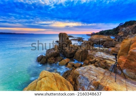 PHoto camera mounted on tripod at scenic sunrise in Tathra beach town facing historic Tathra wharf and colourful rocks.