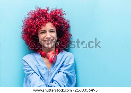 portrait of a smiling young latin woman with a red afro hair in blue background looking at the camera, her red headphones around her neck