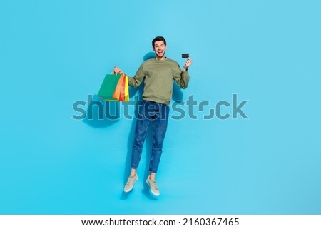 Full size photo of impressed young brunet guy jump with bags card wear shirt jeans shoes isolated on blue background